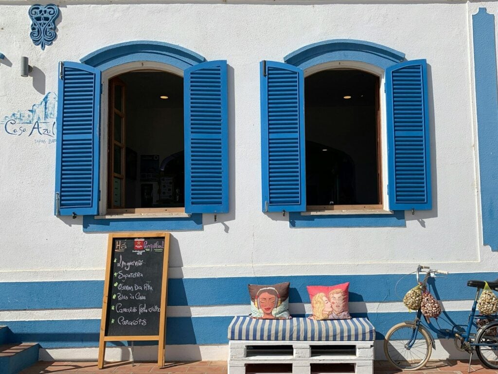 Two multicolored throw pillows on bench near bike parking on white and blue wall showing open blue wooden windows